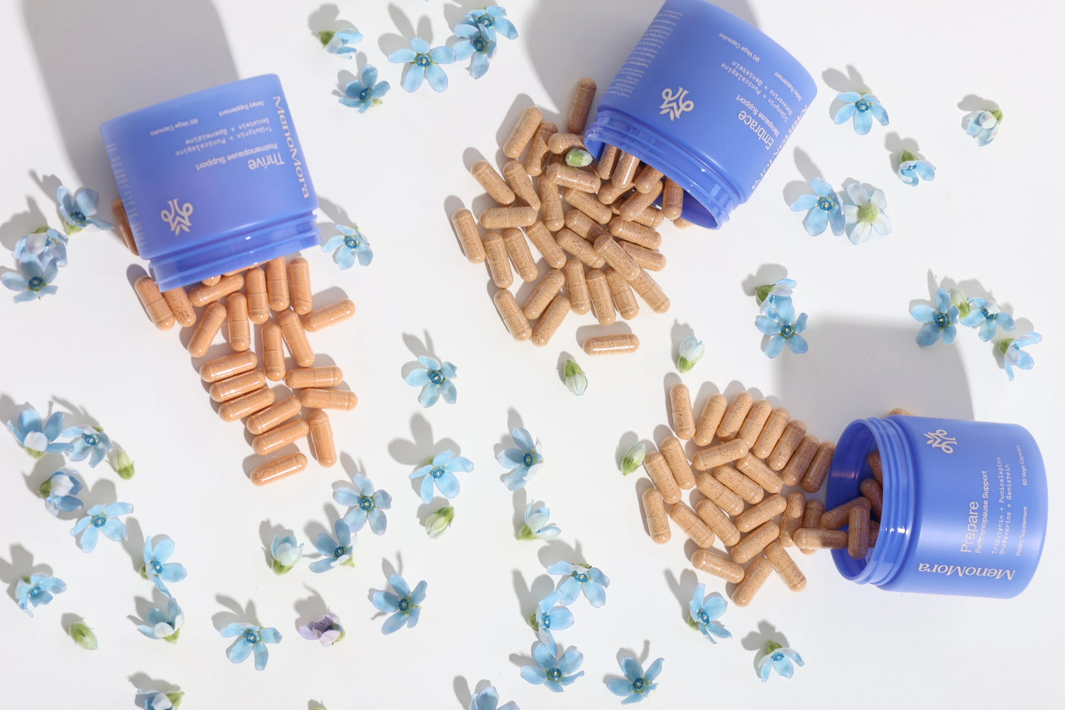 Three blue supplement bottles tipped over with tan capsules spilling out, arranged on a white surface with scattered small blue flowers. The capsules appear to be the product being sold, and the bottles are part of the product presentation rather than separate packaging to exclude.